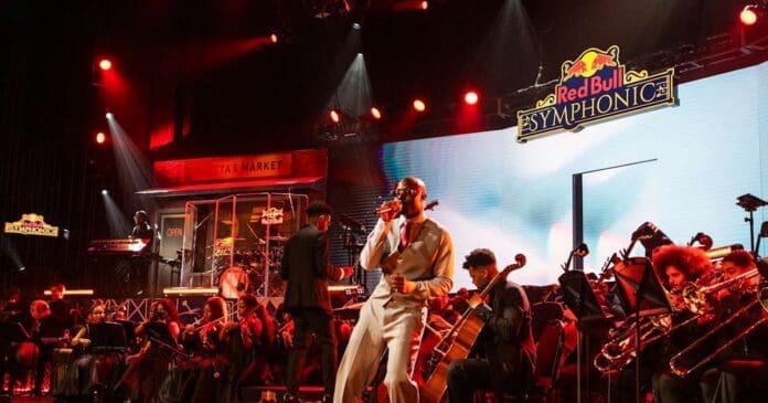 A singer performs on stage with an orchestra during an Asake Red Bull Symphonic event, with musicians playing various instruments under red and yellow stage lighting. Watch Asake stream live for a unique musical experience.