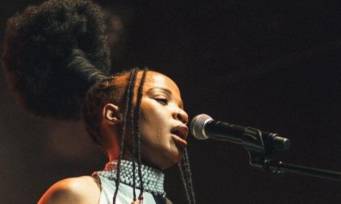 Mozambican singer Assa Matusse, with an elaborate afro hairstyle and beaded necklace, sings passionately into a microphone on stage.