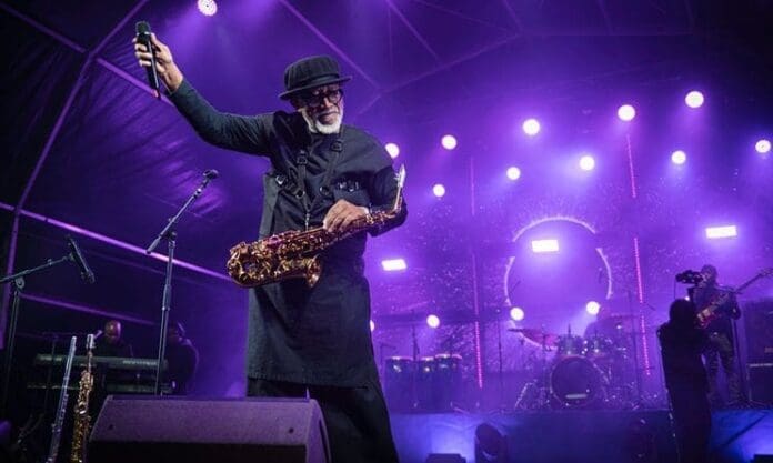 A musician wearing dark clothing and a hat holds a saxophone and a microphone onstage at the Luju Festival in eSwatini, with bright purple stage lights shining and a band performing in the background at this vibrant culture event.