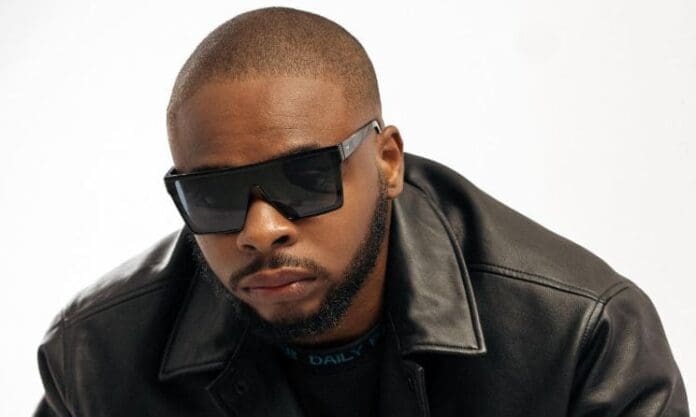 A man with a shaved head wearing dark sunglasses and a black leather jacket stands against a plain white background, channeling DJ Phil’s bold style from the Africa Now DJ Mix on Apple Music.