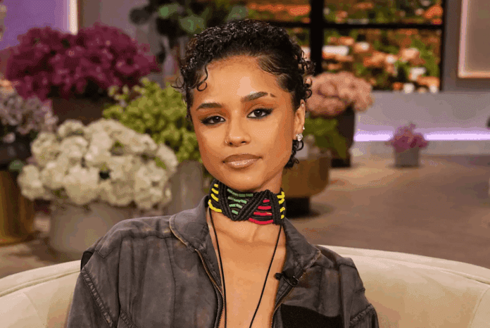 A woman with short curly hair sits indoors, wearing a gray jacket and a colorful choker necklace, with blurred flowers and plants in the background, channeling the vibrant style of US Afrobeats artist Tyla.