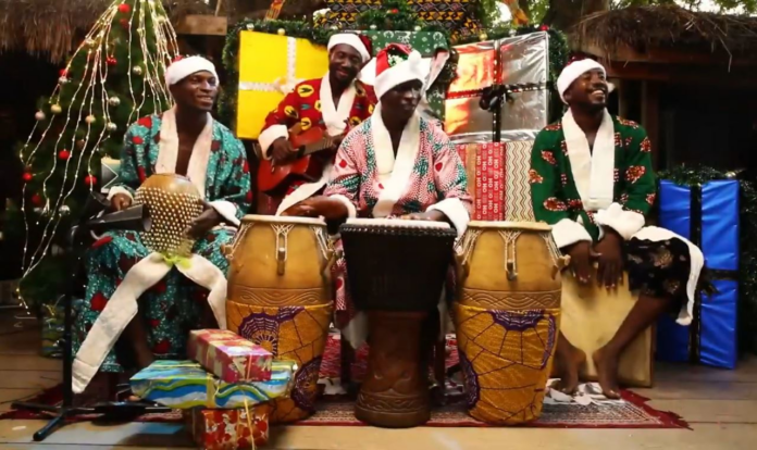 Four men wearing Santa hats and colorful robes play drums and a guitar, surrounded by wrapped gifts and Christmas decorations, celebrating Ghana Christmas music with upbeat Afrobeat rhythms.