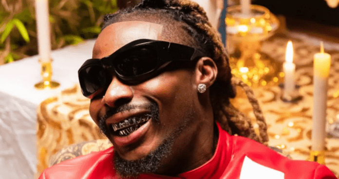 A person with long braided hair, sunglasses, diamond earrings, and grillz smiles while wearing a red jacket. Candles and a decorated table are in the background, reminiscent of the vibrant style seen at an Asake Kenya concert.