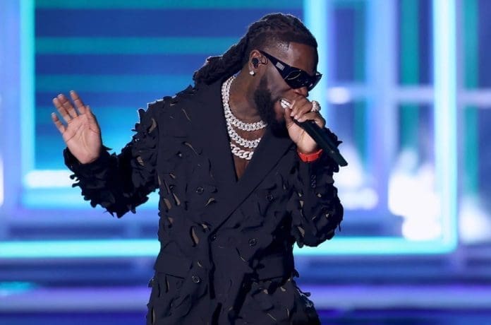 A man wearing a textured black suit, sunglasses, and layered chains sings into a microphone on stage with blue lighting, channeling Burna Boy's style and echoing his African Touring Record for historic revenue.