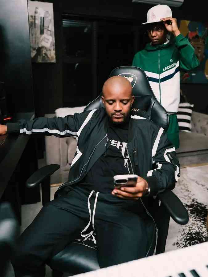 Two men in tracksuits are in a modern room; one sits in a chair looking at his phone, while the other stands behind him adjusting his cap—perhaps discussing the latest Kabza De Small Top Dawg Sessions Mix.