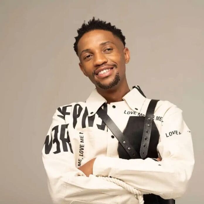 A man stands against a plain background, smiling with his arms crossed, wearing a cream-colored shirt with black text and a black harness—ready to enjoy some Amapiano Mix between friends.