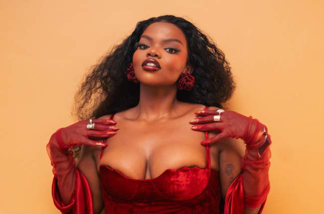 Woman with long curly hair wears a red velvet dress, red gloves, and red rose earrings, posing with her hands near her shoulders against a plain peach background, embodying the vibrant spirit of African music and Amapiano style.
