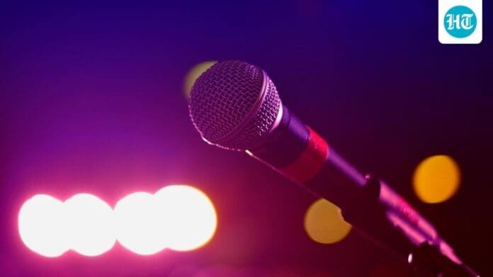A close-up of a microphone in focus with blurred pink and yellow stage lights in the background; an "HT" logo appears in the top right corner, evoking the energy of Nigerian rap and protest music.