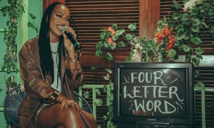 Afro-pop singer Njerae sits on a chair, smiling and holding a microphone next to a TV displaying the words "FOUR LETTER WORD" in white chalk-like text, celebrating the vibrant sounds of Music In Africa.