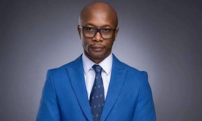 A man wearing glasses, a blue suit, white shirt, and patterned tie stands against a neutral gray background in Lagos, looking directly at the camera—a style often seen at AFRIMA awards and Music In Africa events.