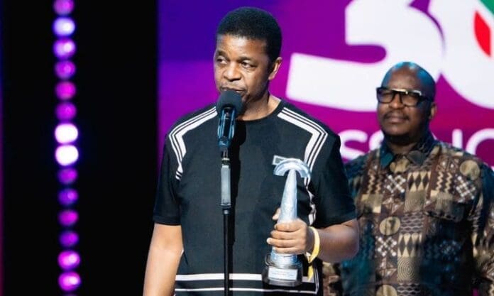 A man holding a silver award speaks into a microphone on stage at the South African Music Awards, with another man standing behind him against a colorful backdrop—highlighting the best in Music In Africa and 2025 entries.