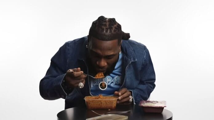 A person with braided hair in a denim jacket eats Jollof rice from a rectangular plastic container with a spoon while sitting at a black table.
