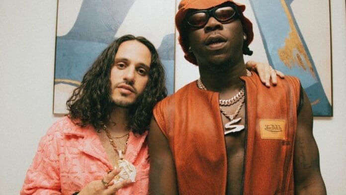 Two men pose together in front of abstract wall art; one rocks a pink outfit and necklace, the other sports an orange vest, sunglasses, and a large chain. This vibrant scene could headline an Unorthodox Reviews feature on Afrobeats style.