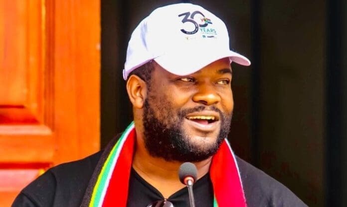 A man wearing a white “30 Years” cap and a colorful scarf speaks into a microphone in front of a wooden door at the 2025 African Music Business Dialogue in Ghana.