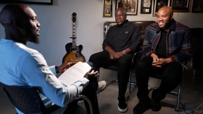 Three men sit indoors having a discussion; one holds a paper, while two others listen. Framed photos, a guitar, and references to Nigerian music executives in the Afrobeats scene are visible in the background.