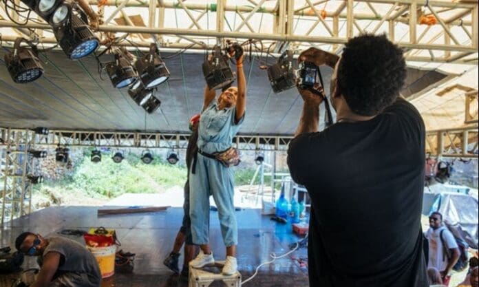 A person stands on a step ladder adjusting stage lights while another, part of the Music In Africa crew, takes a photo; others work nearby on a stage under a metal canopy, contributing to the creative energy in East Africa.