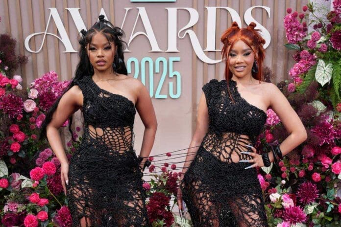 Two women from TxC stand side by side in matching black, netted dresses at a BET award event, with colorful flowers and a sign reading "AWARDS 2025" in the background, celebrating Amapiano music’s growing influence.