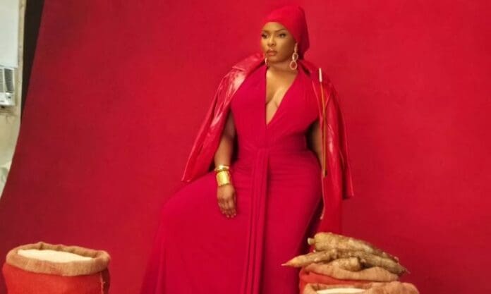 A woman dressed in a vibrant red outfit and headscarf poses against a red backdrop, with burlap sacks of tubers at her feet—a striking scene reflecting Zanzibar’s rich culture, reminiscent of visuals from the Trace Awards 2025.