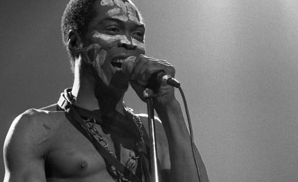 A shirtless man with face paint sings into a microphone on stage under dramatic lighting, channeling the energy of Fela and the spirit of an African Giant from Nigeria.