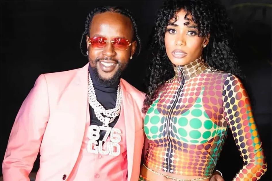 A man in a pink suit and sunglasses stands next to Toni-Ann Singh, who wears a colorful, patterned outfit. Both are facing the camera and posing confidently against a dark background.