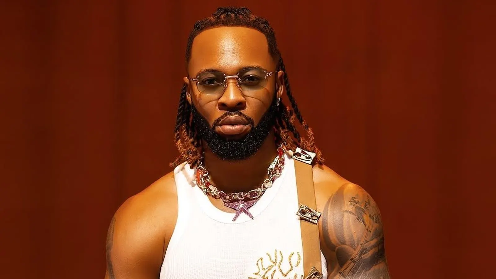 A man with sunglasses, braided hair, a trimmed beard, and tattoos on his arm wears a white tank top and layered necklaces, channeling Afrobeats style as he stands against a plain brown background.