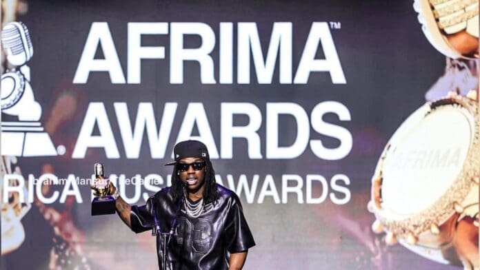 A person in black clothing and sunglasses holds a trophy while speaking on stage at the AFRIMA Awards, embodying Continental Excellence, with a large event logo displayed in the background.