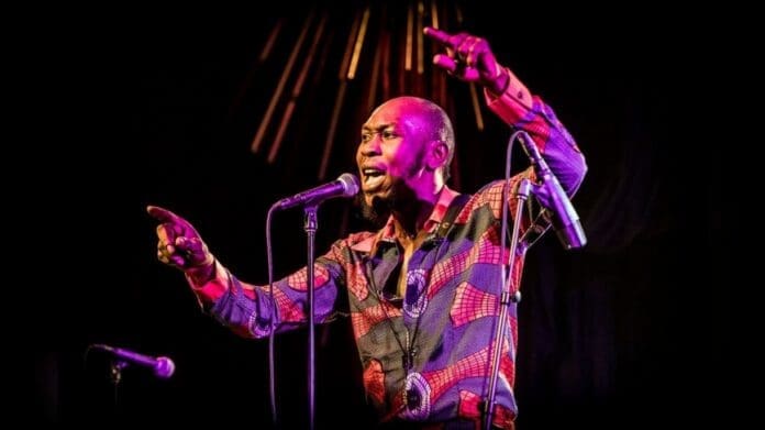 A man in a patterned shirt, reminiscent of Fela’s vibrant style, sings passionately into a microphone on stage, gesturing with both hands under dramatic lighting.