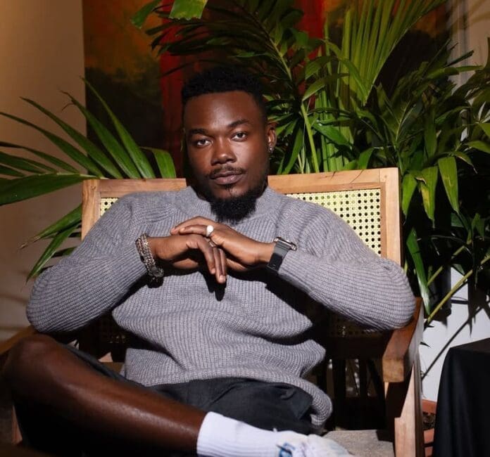 A man in a grey sweater sits in a wooden chair with greenery in the background, looking directly at the camera with his hands clasped—channeling the cool confidence of Camidoh, known for his Rides Solo vibes on 3Music.