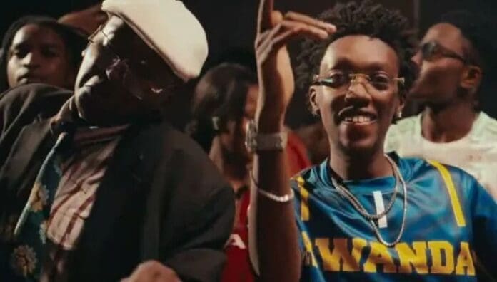 A group of people stand together; a man in glasses and a Rwanda jersey smiles and gestures, while another man beside him wears a hat and jacket, capturing the spirited vibe of East African Songs featured by OkayAfrica in August 2025.