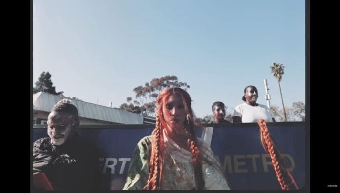 A person with long orange braids stands in front of three others near a blue METRO sign, outdoors on a clear day, capturing a moment that could inspire the 2025 OkayAfrica list of top Afro Jazz Songs.