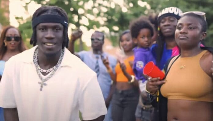 A group of people stand outdoors; a man in front wears a white shirt and headband, while a woman beside him holds a red cup. Trees are visible in the background, capturing an OkayAfrica gathering with West African songs on a September 2025 afternoon.