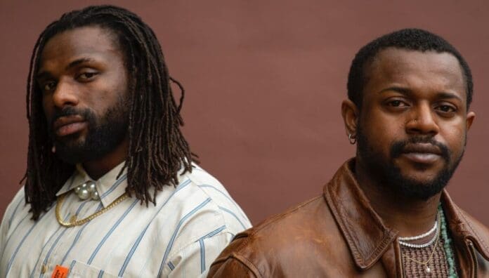 Two men stand side by side in front of a brown background. One wears a striped shirt with dreadlocks; the other sports a brown leather jacket and chain necklace, capturing the vibrant style often seen in OkayAfrica’s top songs features.
