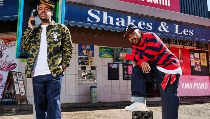 Two men pose in front of a storefront named "Shakes & Les"; one stands holding a payphone while the other crouches on a ledge, capturing the vibe of amapiano songs and the energy of the upcoming 2025 music scene.
