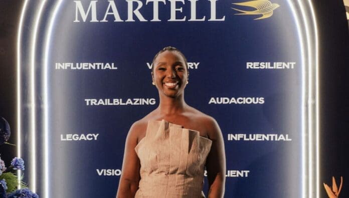 A woman in a beige, off-shoulder dress stands smiling in front of a blue Martell promotional backdrop with motivational words, celebrating global growth and Afrobeats alongside Sheniece Charway.