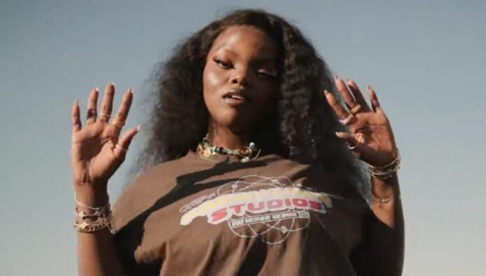 A person with long, wavy hair stands outdoors, wearing a brown graphic t-shirt and jewelry, both hands raised and fingers spread against a clear sky—channeling the bold style of new artists shaping Amapiano in 2025.