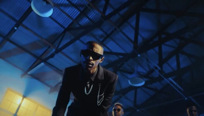 A man in a black suit and sunglasses leans forward under industrial ceiling lights in a dimly lit room with blue lighting, lost in the rhythms of Amapiano songs set to stream everywhere by October 2025.