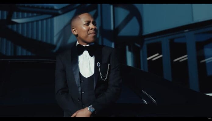 A person in a black tuxedo with a bow tie and chain accessory stands beside a black car in front of a modern building, exuding the style often celebrated by OkayAfrica’s Top Songs.