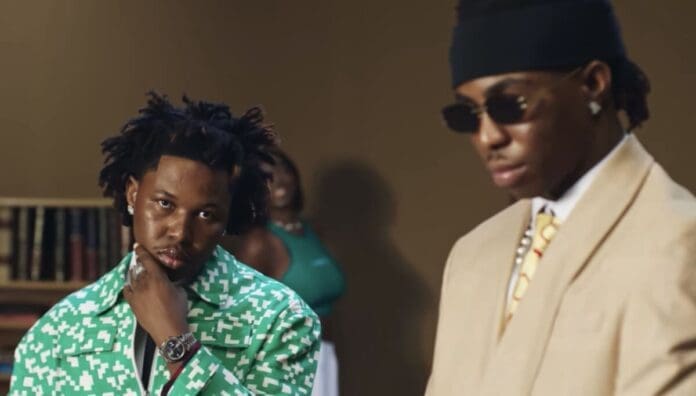 Two men pose indoors; one in a patterned green jacket touches his chin, while the other sports a beige suit and sunglasses. A blurred figure in green stands behind them—an image with the bold style seen in OkayAfrica’s coverage of African songs.
