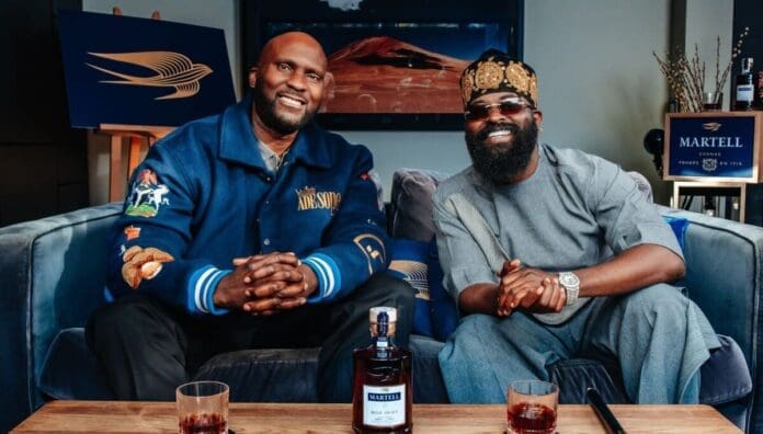Two men sit on a couch with smiles, facing the camera; a bottle of Martell Blue Swift and two glasses are on the table, set for Swift Conversations about King Smade and Afrobeats.