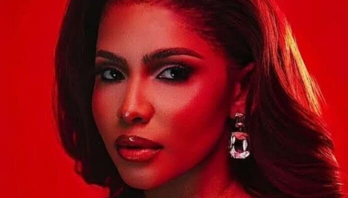 A woman with long, styled hair and glossy lipstick poses in front of a red background, wearing large, dangling earrings—her look channeling the vibrant energy of the best Amapiano songs featured by OkayAfrica.