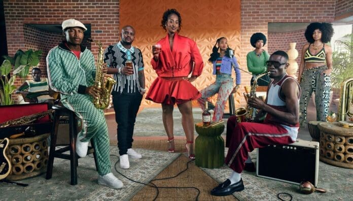 A group of nine people pose indoors with musical instruments and drinks, featuring bold, colorful outfits and a retro-styled setting with plants and brick walls, channeling the vibrant energy of OkayAfrica.