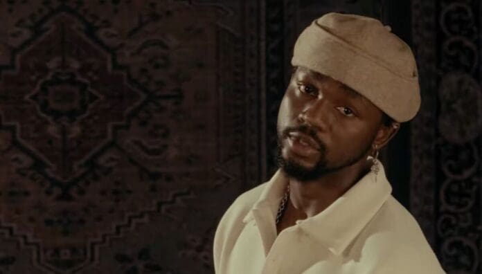 A man with a beard and earring, wearing a beige hat and cream shirt, stands in front of a dark patterned tapestry, evoking the vibrant energy of West African songs celebrated by OkayAfrica.