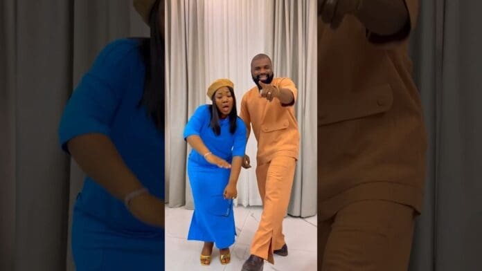 A woman in a blue dress and a man in an orange outfit stand indoors, both posing energetically in front of white curtains, radiating exceeding joy as if celebrating a lively praise anthem.
