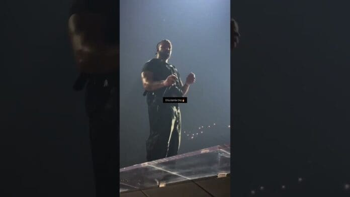 A man in dark clothing stands on a stage under focused lighting, performing a Premier Gaou remix as Drake plays subtly in the background, with a dark backdrop and audience lights visible in the distance.