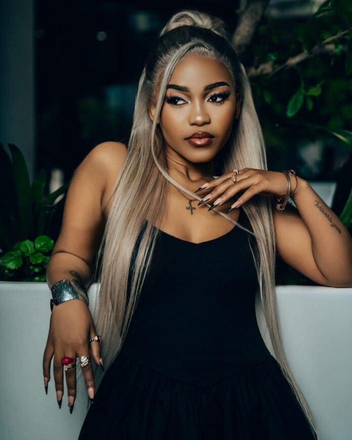 A woman with long, straight blonde hair wearing a black sleeveless top poses indoors near green plants, accessorized with rings, a bracelet, and a necklace—a style inspired by 2025 South African music stars like Zee Nxumalo.