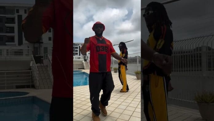 Two people stand near a swimming pool, one in a red and black outfit and another in yellow and black, discussing an upcoming music release with buildings and a cloudy sky in the background.