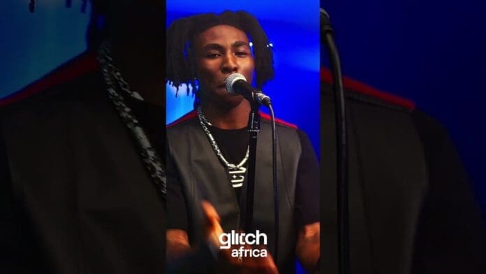 A man sings into a microphone on stage, wearing a black shirt, vest, and chain necklace during Glitch Sessions. The text "glitch africa" is displayed at the bottom of the image.
