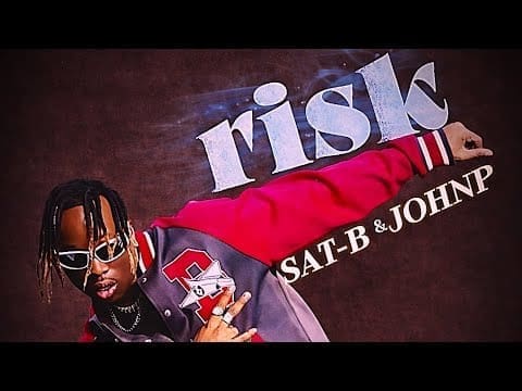 A man in sunglasses and a red varsity jacket poses in front of the word "risk" and the names "SAT-B & JOHNP," hinting at the official video for "Risk SAT-B" by Johnp artist on a dark background.
