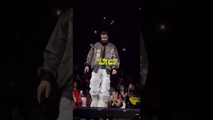 A man in a jacket and white pants stands on stage during a concert, with audience phone lights in the background. Text on screen reads, "All the girls that's here," referencing Drake’s Afro lyrics from the One Remix.