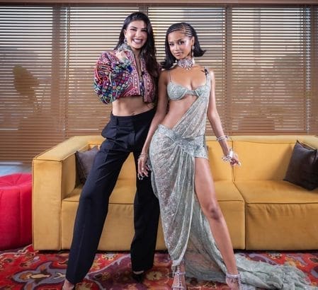 Two women pose indoors; one resembles Jacqueline Fernandez in a colorful jacket and black pants, while the other channels Tyla in a shimmering, draped outfit with a high slit. They stand before a yellow couch and window blinds.